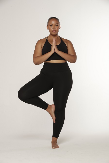 Woman doing yoga tree pose with hands to heart wearing black sports bra and black ankle biter leggings.